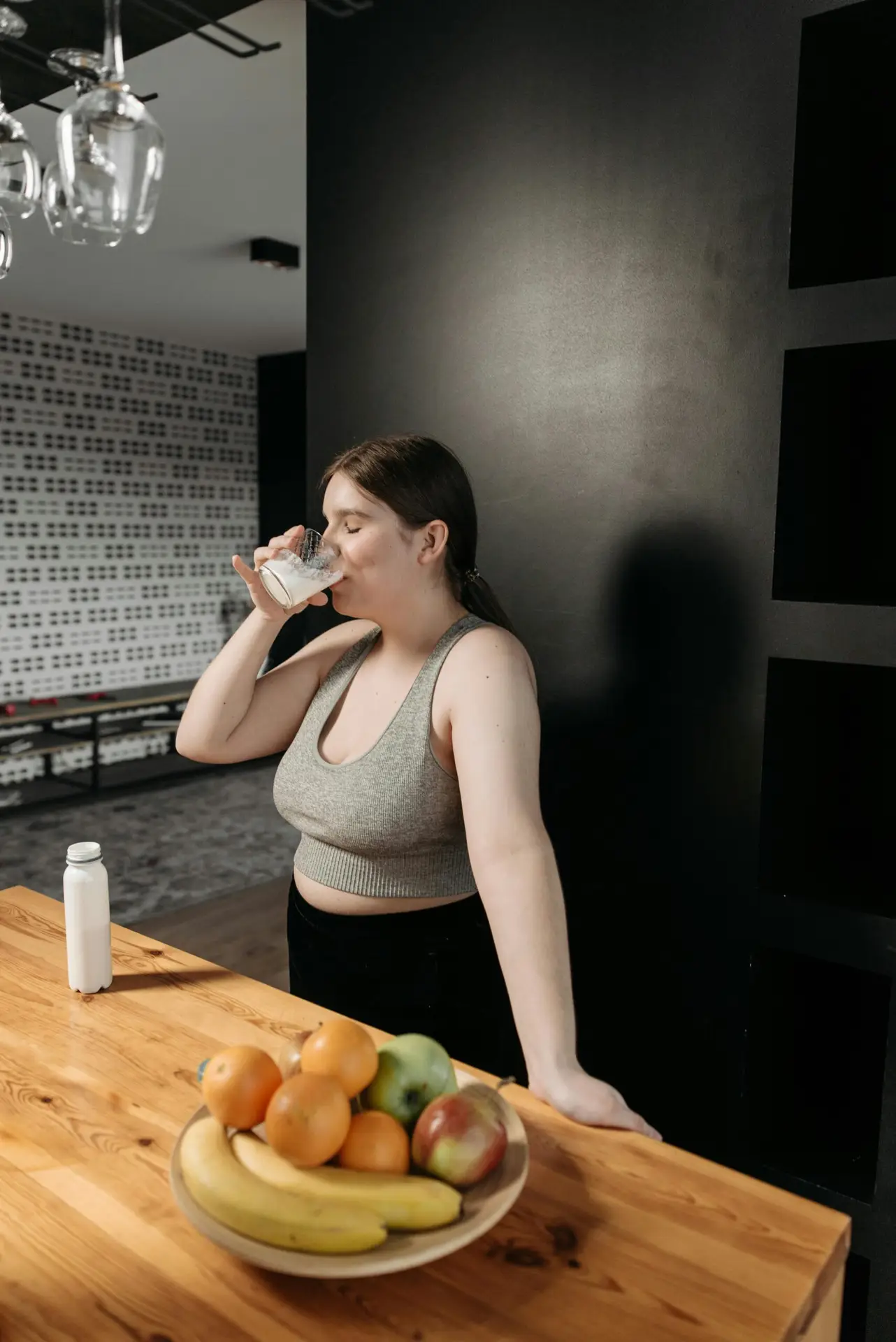 Woman in a gray tank top drinking water beside a table with fruits
